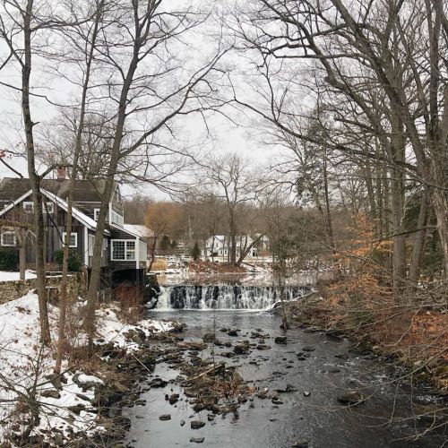 A serene winter scene featuring a house by a river with a small waterfall, surrounded by bare trees and snow.