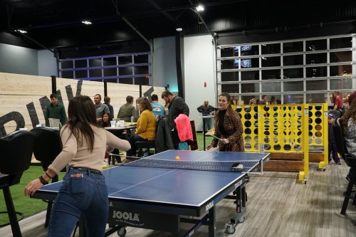 Two women play table tennis in a lively indoor space with people seated at tables in the background.
