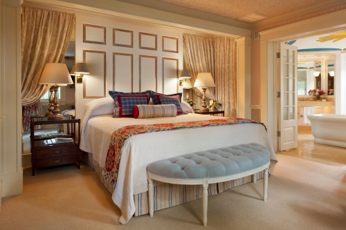 Cozy bedroom featuring a large bed with decorative pillows, a bench, and elegant curtains, with a bathroom visible in the background.