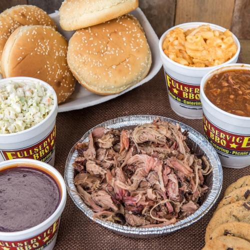 A spread of BBQ pulled pork, coleslaw, baked beans, macaroni and cheese, and sesame seed buns on a rustic table.