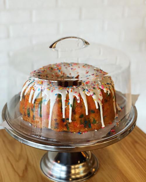 A colorful bundt cake with icing and sprinkles, displayed under a glass dome on a wooden table.