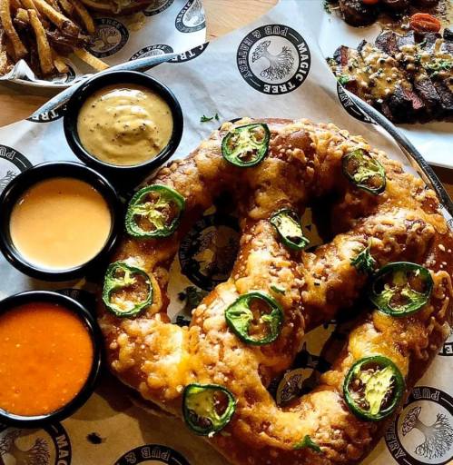 A large, golden-brown pretzel topped with jalapeños, served with three dipping sauces in black bowls.