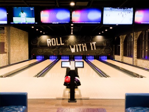 A bowling alley with lanes, screens showing sports, and a sign that says "ROLL WITH IT." Colorful bowling balls are visible.