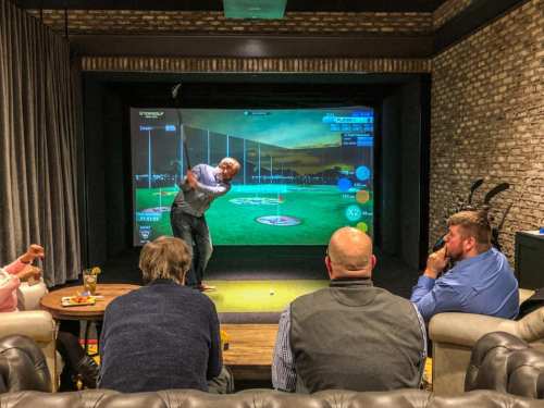 A group of people watching a golfer using a virtual golf simulator in a cozy indoor setting.