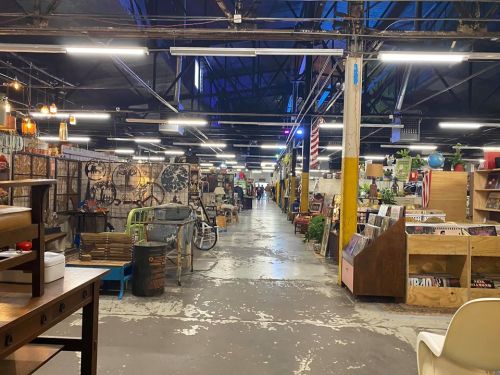 A spacious vintage market with various booths, showcasing antiques and collectibles under industrial lighting.