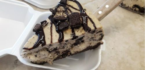 A slice of cookie dough cheesecake topped with chocolate drizzle and crushed cookies, served in a takeout container.