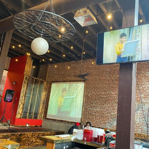A cozy restaurant interior with exposed brick walls, featuring a TV displaying a painting tutorial and warm lighting.