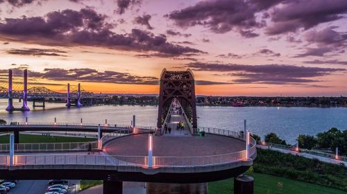 A scenic view of a bridge at sunset, with clouds reflecting colors over a river and surrounding greenery.