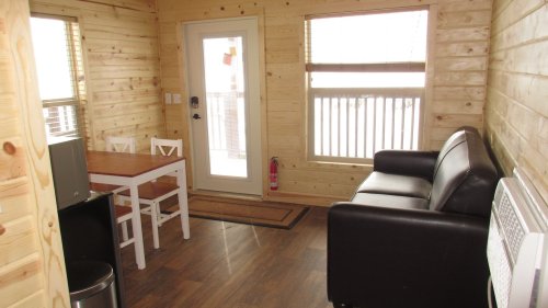 Cozy wooden interior of a cabin with a dining table, black sofa, and large windows letting in natural light.