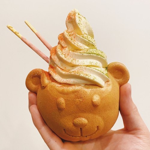 A hand holding a bear-shaped dessert filled with soft serve ice cream, topped with colorful sprinkles and two sticks.