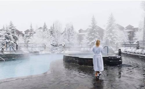 A person in a robe walks towards a steaming hot tub surrounded by snow-covered trees and a winter landscape.