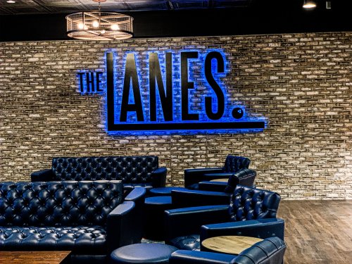 Interior of a lounge with blue leather seating and a brick wall featuring the illuminated sign "THE LANES."