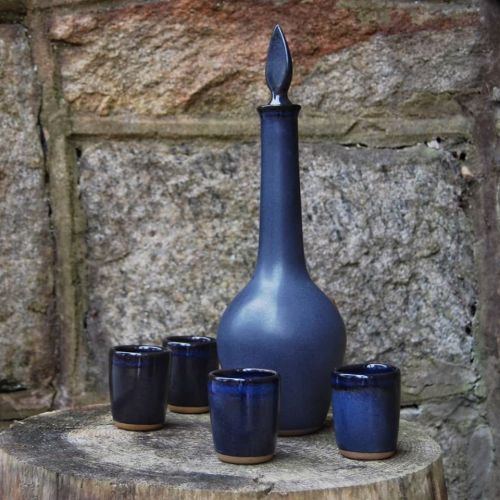 A dark blue ceramic bottle with a pointed lid, surrounded by four matching small cups, set against a stone wall.