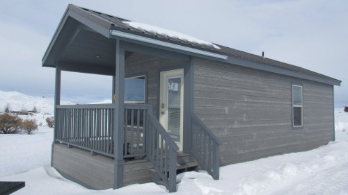 A small gray house with a porch, surrounded by snow and mountains under a cloudy sky.