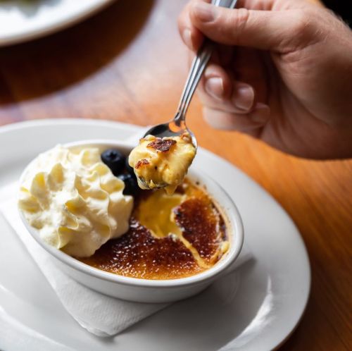 A hand holding a spoon scoops crème brûlée topped with whipped cream and a blueberry, served in a small dish.