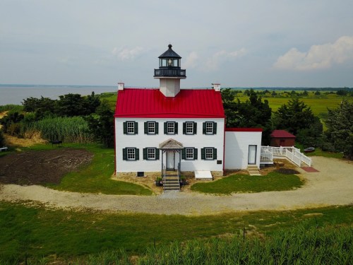 A white lighthouse with a red roof, surrounded by greenery and a calm body of water in the background.