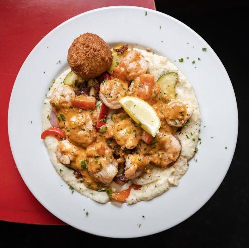 A plate of shrimp and grits topped with a creamy sauce, garnished with lemon and herbs, alongside a fried ball.