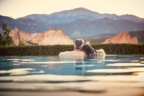 A couple relaxes in a pool, embracing with mountains in the background during sunset.