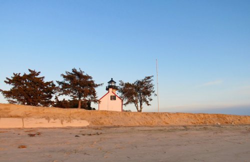 A pink lighthouse stands near the beach, surrounded by trees under a clear blue sky at sunset.