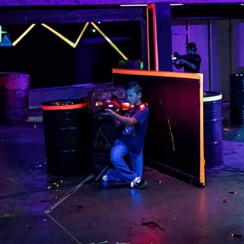 A child crouches behind a barrier, aiming a laser tag gun in a colorful, neon-lit arena.
