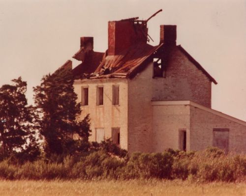 Abandoned two-story house with a damaged roof and boarded windows, surrounded by overgrown grass and trees.