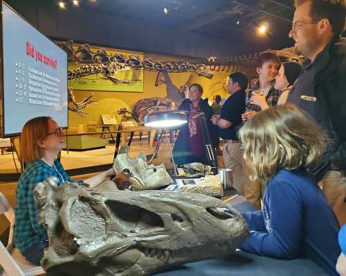 A group of people engages with a dinosaur exhibit, examining fossils and a presentation screen in a museum setting.