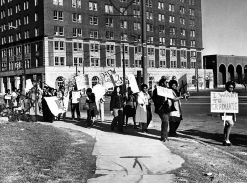 Black and white photo of a group of protesters holding signs, marching in a city street. Buildings in the background.