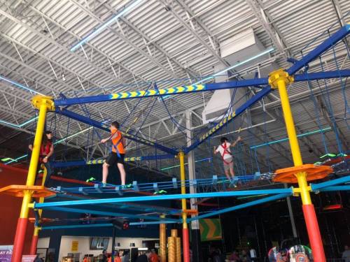 Two children navigate a colorful indoor ropes course, suspended above a play area with bright decorations.