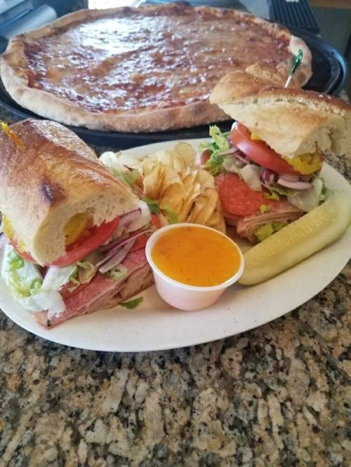 A plate with a sandwich, chips, a pickle, and dipping sauce, with a pizza in the background.