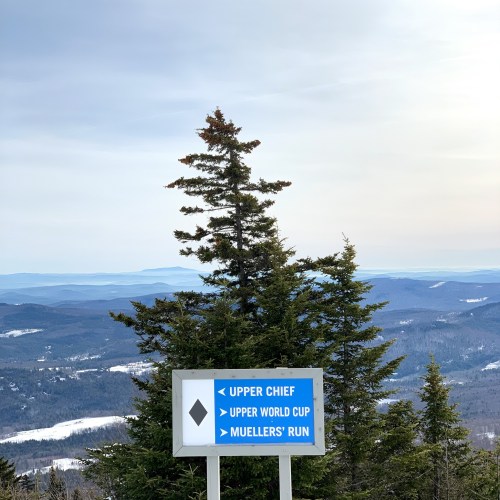 Signpost indicating ski trails: Upper Chief, Upper World Cup, and Mueller's Run, with a scenic mountain view in the background.