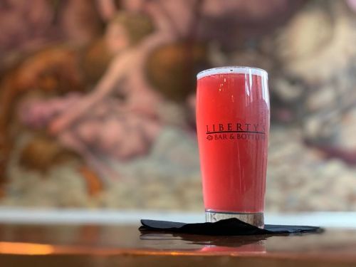 A vibrant pink drink in a glass with "Liberty's Bar & Bottles" logo, set against a blurred artistic background.