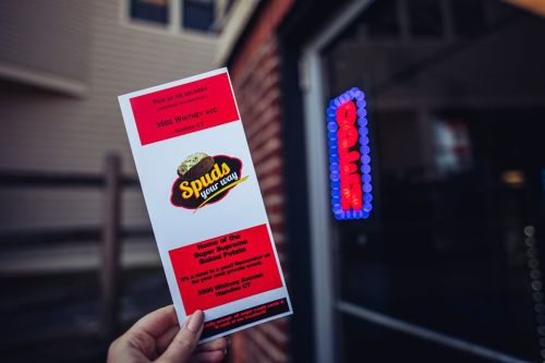 A hand holds a menu for "Spuds Your Way," featuring baked potatoes, outside a restaurant with a neon sign.