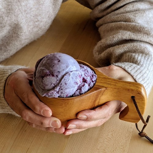 A person holds a wooden bowl filled with purple ice cream, resting on a wooden table.