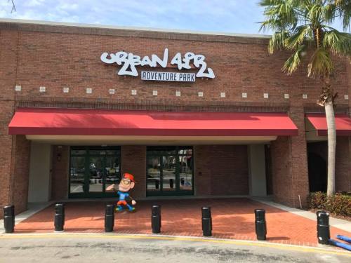 Entrance of Urban Air Adventure Park with a cartoon character in front and palm trees nearby. Red awnings above the doors.
