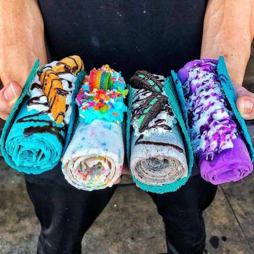 A person holds four colorful rolled desserts, each with unique toppings and vibrant blue tortillas.