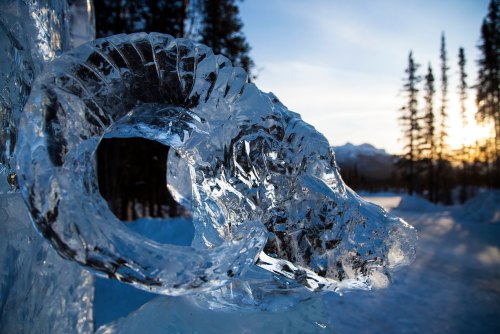 A close-up of an intricately carved ice sculpture of a ram's head, with trees and mountains in the background at sunset.