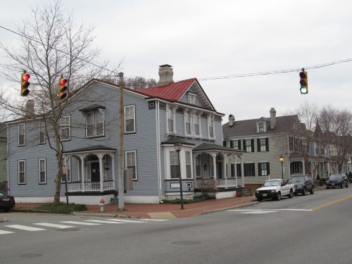 A street view of a blue house with a red roof, traffic lights, and parked cars in a residential area.