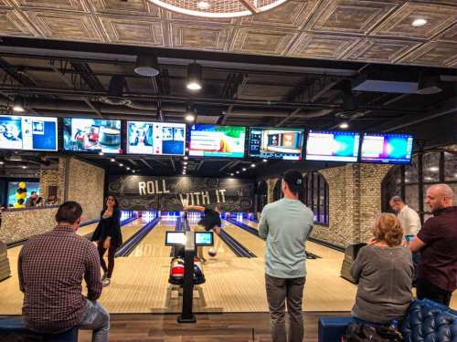 A bowling alley with multiple screens, featuring people watching and preparing to bowl. The atmosphere is lively and modern.