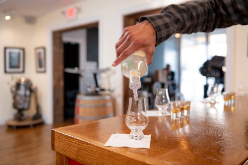 A hand pours a clear liquid from a bottle into a tasting glass on a wooden bar, with distillery equipment in the background.