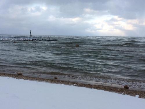 Waves gently lap at a snowy beach under a cloudy sky, with a distant pier visible on the horizon.