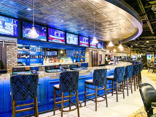 A modern bar with blue accents, high stools, multiple TVs, and a well-stocked shelf of liquor in a stylish setting.