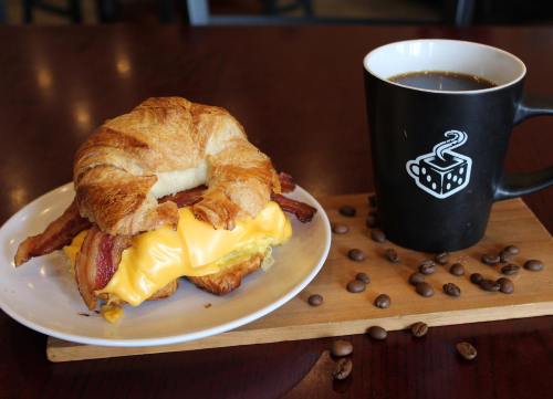 A croissant sandwich with bacon and melted cheese on a plate, next to a coffee cup and scattered coffee beans.