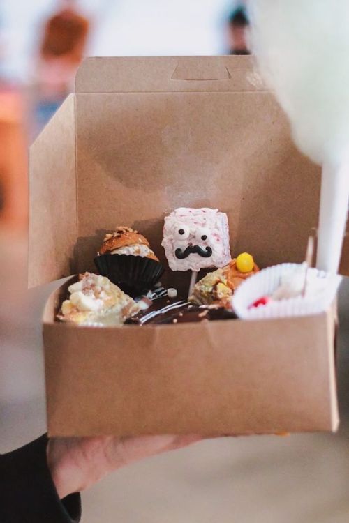 A hand holding a takeout box filled with assorted desserts, including a cupcake, a treat with a mustache, and cotton candy.