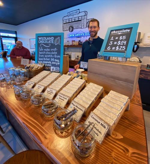 A man stands behind a wooden counter displaying various chocolate bars and prices at a cozy café.