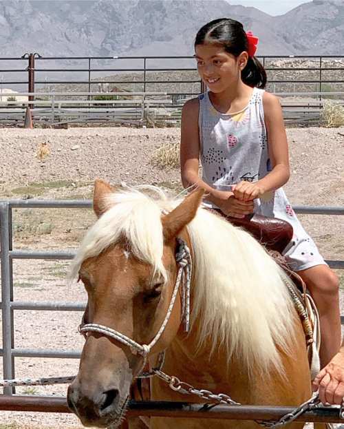 A young girl with a red bow smiles while riding a light brown horse in a sunny outdoor setting.