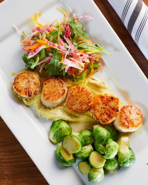 Grilled scallops on a plate with salad and Brussels sprouts, garnished with colorful shredded vegetables.