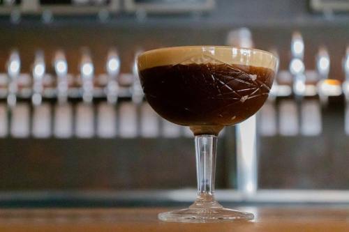 A stylish cocktail in a crystal glass, featuring a dark liquid topped with a creamy foam, set against a blurred bar background.