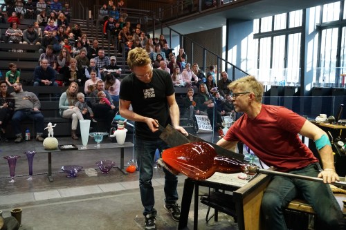 Two glassblowers demonstrate their craft in front of an audience, showcasing colorful glass pieces and tools.