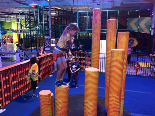 A child in a harness jumps between colorful pillars in a vibrant indoor play area, with other kids nearby.