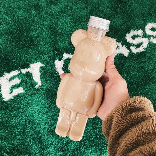 A hand holding a bear-shaped bottle filled with a creamy beverage, set against a green textured background.